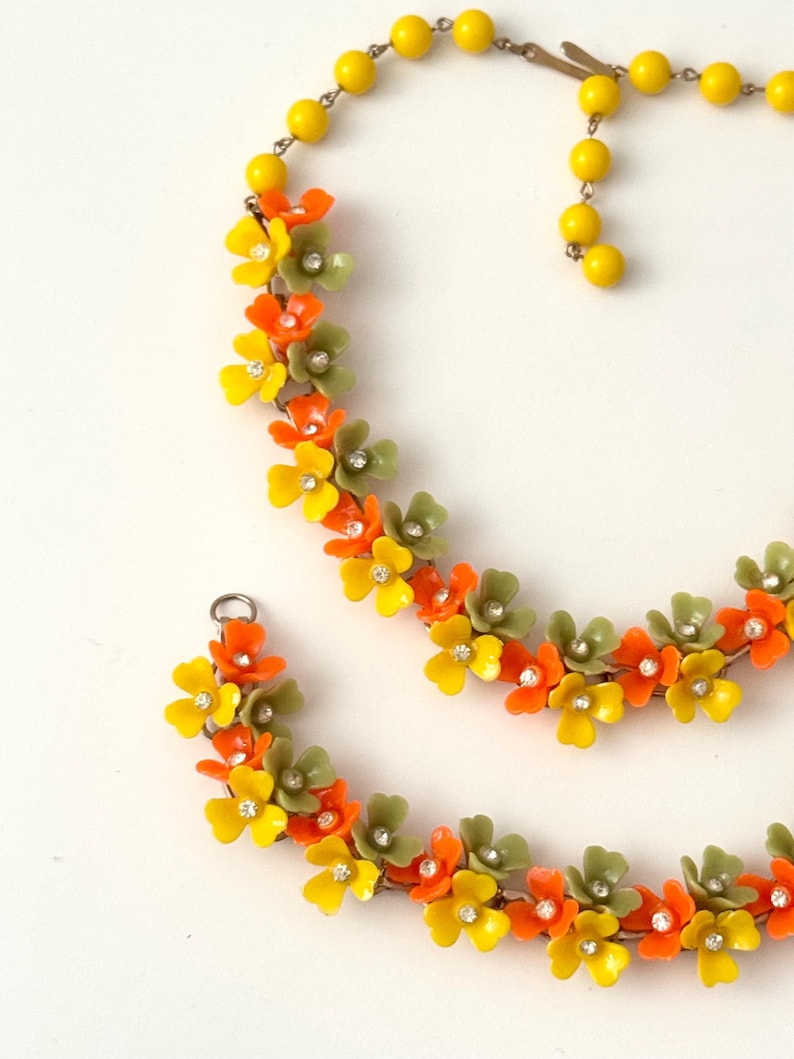 May include: A necklace and bracelet set featuring a series of small, five-petaled flowers in yellow, orange, and olive green. Each flower is accented with a small, clear rhinestone at the center. The necklace includes a strand of yellow beads.