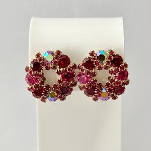 Puede incluir: Un par de pendientes circulares con un grupo de cristales facetados de color rojo intenso engastados en metal dorado. Cada pendiente tiene un diseño en forma de corona con cristales iridiscentes intercalados que reflejan la luz.