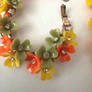 May include: A link bracelet featuring a series of three-petaled flowers in shades of olive green, bright orange, and yellow. Each flower is centered with a small, clear, round rhinestone. The bracelet is constructed with gold-tone metal links.