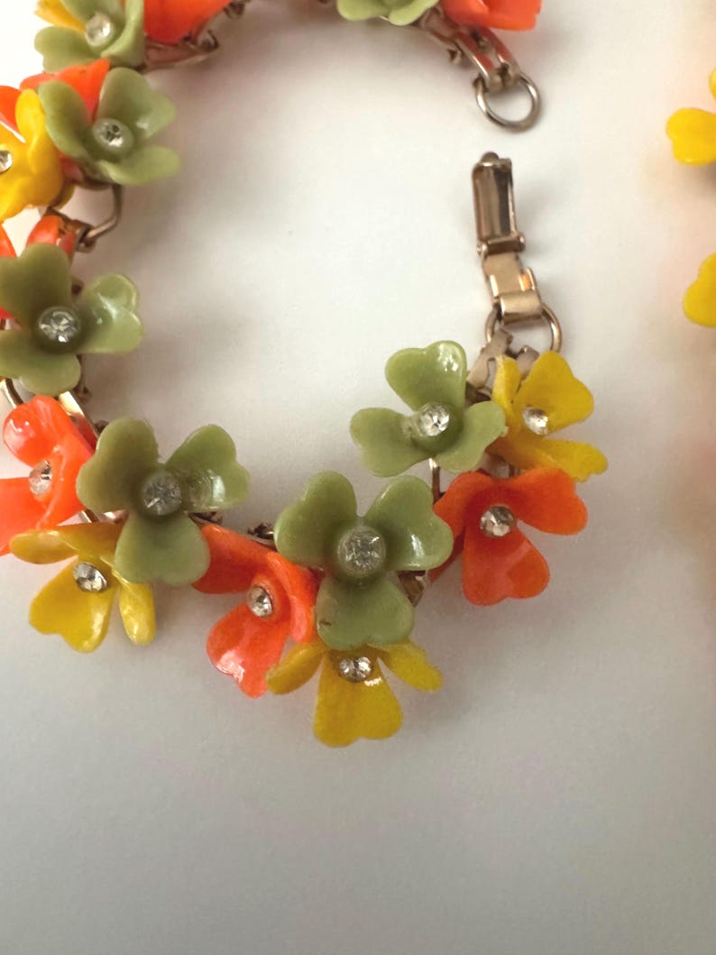 May include: A link bracelet featuring a series of three-petaled flowers in shades of olive green, bright orange, and yellow. Each flower is centered with a small, clear, round rhinestone. The bracelet is constructed with gold-tone metal links.