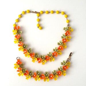May include: A jewelry set consisting of a necklace and a bracelet featuring yellow, orange, and sage green plastic flowers. Each flower has a small clear rhinestone at its center. The necklace includes a strand of yellow beads and a metal clasp.