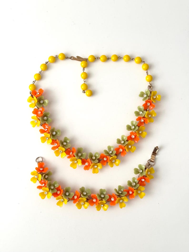 May include: A jewelry set consisting of a necklace and a bracelet featuring yellow, orange, and sage green plastic flowers. Each flower has a small clear rhinestone at its center. The necklace includes a strand of yellow beads and a metal clasp.