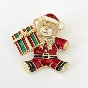 May include: A gold-tone metal brooch shaped like a teddy bear wearing a red Santa suit with a white-trimmed hat. The bear holds a rectangular gift box with red and green stripes. The brooch features red and white enamel and clear rhinestones.