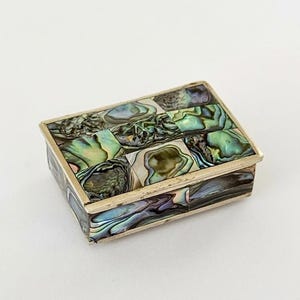 May include: A rectangular box featuring a metallic frame and an exterior surface inlaid with iridescent abalone shell tiles. The shell pieces display a variety of green, blue, and purple tones with a natural, swirling, and reflective pearlescent finish.
