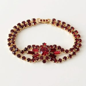 Könnte beinhalten: Ein goldfarbenes Armband mit zwei Reihen aus runden, tiefroten Strasssteinen. Der Mittelpunkt ist ein komplexes Arrangement aus runden und baguetteförmigen roten Strasssteinen in Sternform. Das Armband verfügt über einen sicheren Metallverschluss.