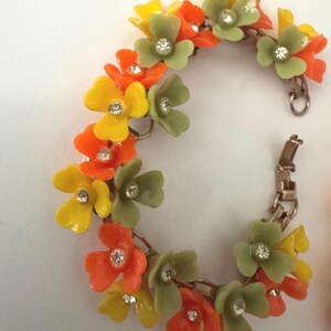 May include: A link bracelet featuring a series of connected floral charms in shades of yellow, orange, and sage green. Each flower has a small, clear, round rhinestone set in the center. The bracelet includes a metallic clasp mechanism for closure.