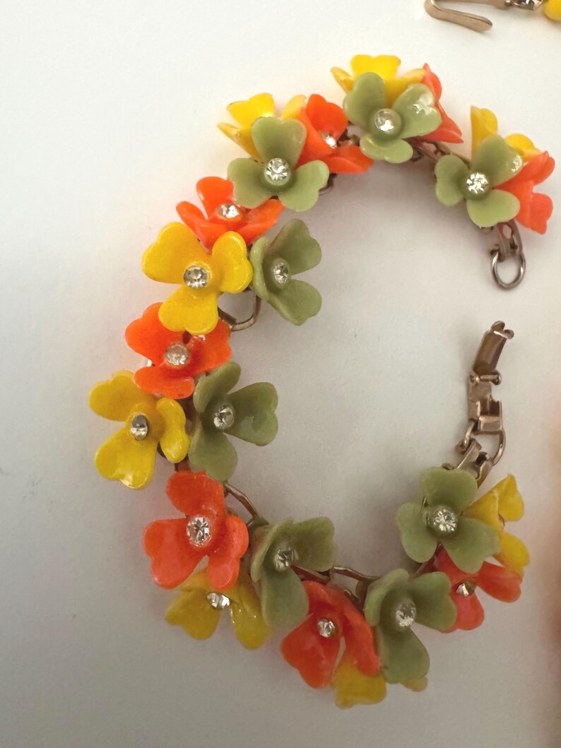 May include: A link bracelet featuring a series of connected floral charms in shades of yellow, orange, and sage green. Each flower has a small, clear, round rhinestone set in the center. The bracelet includes a metallic clasp mechanism for closure.