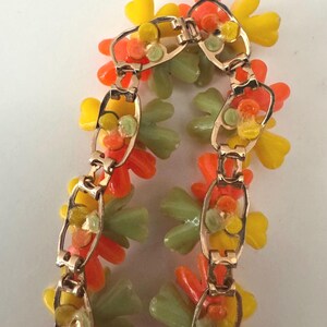 May include: A gold-tone metal link bracelet featuring clusters of translucent plastic flowers in shades of bright orange, sunny yellow, and pale sage green. The bracelet is constructed with repeating oval-shaped metal links that connect the floral elements.