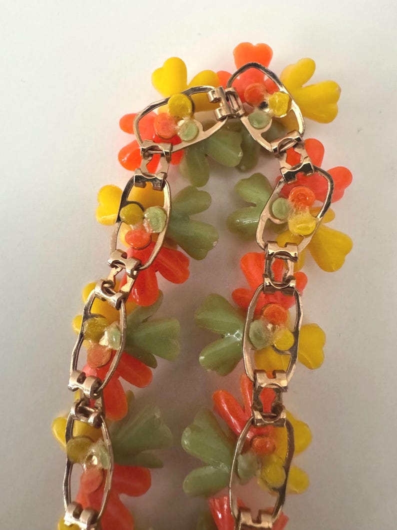 May include: A gold-tone metal link bracelet featuring clusters of translucent plastic flowers in shades of bright orange, sunny yellow, and pale sage green. The bracelet is constructed with repeating oval-shaped metal links that connect the floral elements.