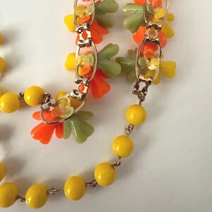 May include: A necklace featuring a gold-tone metal chain interspersed with bright yellow spherical beads and clusters of small, translucent orange and pale green flower-shaped charms attached to the links of the chain.