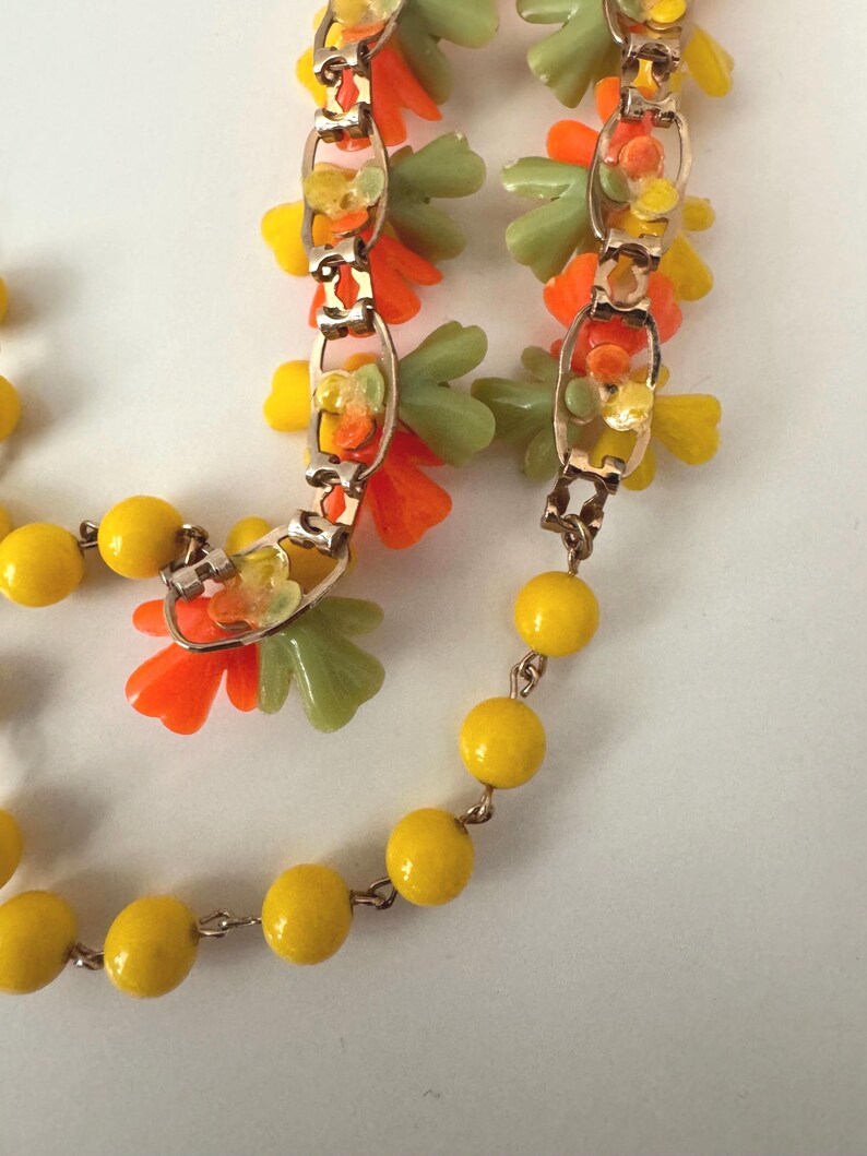 May include: A necklace featuring a gold-tone metal chain interspersed with bright yellow spherical beads and clusters of small, translucent orange and pale green flower-shaped charms attached to the links of the chain.