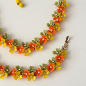 May include: A necklace and bracelet set featuring a floral design with small yellow, orange, and sage green petals. Each flower is accented with a clear rhinestone at the center. The necklace includes a strand of yellow beads and a metal clasp.