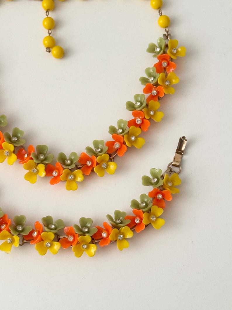 May include: A necklace and bracelet set featuring a floral design with small yellow, orange, and sage green petals. Each flower is accented with a clear rhinestone at the center. The necklace includes a strand of yellow beads and a metal clasp.
