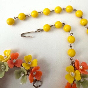 May include: A necklace featuring a strand of bright yellow beads connected to a cluster of multi-colored floral charms. The flowers are yellow, orange, and pale green, each centered with a small clear rhinestone. A silver-toned metal hook clasp.