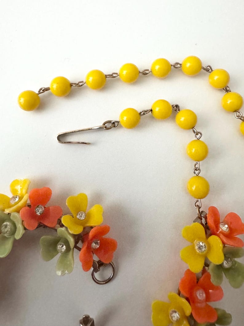 May include: A necklace featuring a strand of bright yellow beads connected to a cluster of multi-colored floral charms. The flowers are yellow, orange, and pale green, each centered with a small clear rhinestone. A silver-toned metal hook clasp.