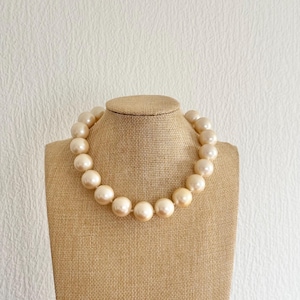 Può includere: Una collana di perle color crema esposta su un busto di gioielli beige. La collana presenta grandi perle rotonde, creando un look classico ed elegante. Le perle sono uniformemente distanziate e hanno una lucentezza sottile.