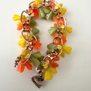 May include: A gold-tone metal link bracelet featuring clusters of small, molded plastic flower-shaped charms in vibrant shades of yellow, orange, and sage green. The bracelet is constructed with rectangular metal links and a secure fold-over clasp.