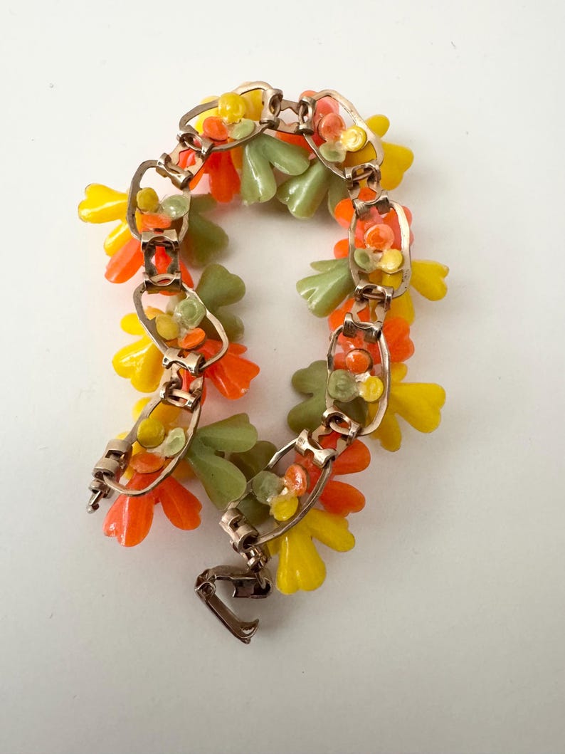 May include: A gold-tone metal link bracelet featuring clusters of small, molded plastic flower-shaped charms in vibrant shades of yellow, orange, and sage green. The bracelet is constructed with rectangular metal links and a secure fold-over clasp.