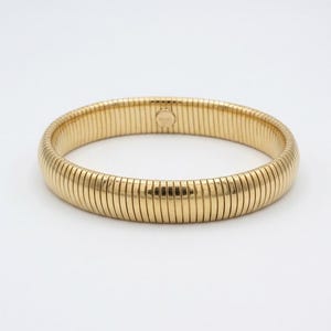 Könnte beinhalten: Ein goldfarbenes Metallarmband mit einem flexiblen, dehnbaren Design, das aus zahlreichen vertikalen, rechteckigen Gliedern besteht. Die Oberfläche hat ein poliertes, reflektierendes Finish und ein kleiner runder Stempel ist auf dem Innenband sichtbar.