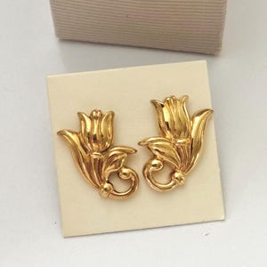 Op de afbeelding: Een paar goudkleurige metalen oorbellen in de vorm van gestileerde tulpen met gebogen stelen en bladeren. De oorbellen zijn bevestigd op een vierkante, gebroken witte displaykaart, geplaatst voor een open kartonnen doosje.