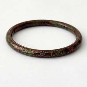 Könnte beinhalten: Ein runder Armreif mit dunkelbrauner Basis und aufwendiger Cloisonné-Emaille-Arbeit. Die Oberfläche ist mit einem sich wiederholenden Blumenmuster in Grün-, Rot- und Goldtönen verziert, die durch feine Metalldrahtkonturen getrennt sind.