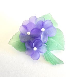 Könnte beinhalten: Blumenbrosche mit drei lila Blüten mit weißen Zentren und grünen Blättern. Die Brosche besteht aus einem durchscheinenden Material, das ein zartes und elegantes Aussehen erzeugt. Die Brosche ist etwa 5 cm lang.