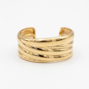 May include: A polished gold-tone cuff bracelet featuring a wide, open-ended band design. The surface is composed of three smooth, rounded, parallel ridges that wrap around the wrist, creating a layered appearance with a high-shine finish.