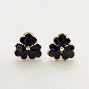 May include: A pair of stud earrings featuring a three-petaled flower design. Each earring has a glossy black enamel finish encased in a gold-toned metal border, with a small gold-toned metal sphere centred at the heart of the flower petals.