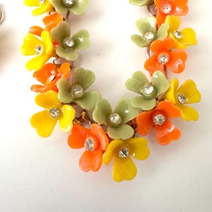 May include: A floral necklace featuring a series of connected blossoms in shades of bright yellow, vibrant orange, and muted sage green. Each individual flower is accented with a small, clear, sparkling rhinestone set in the center of the petals.
