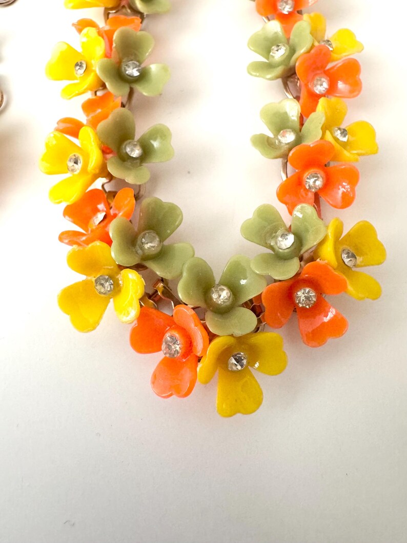 May include: A floral necklace featuring a series of connected blossoms in shades of bright yellow, vibrant orange, and muted sage green. Each individual flower is accented with a small, clear, sparkling rhinestone set in the center of the petals.