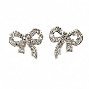 May include: A pair of silver-toned stud earrings shaped like bows. Each bow is crafted from a polished metal frame and is encrusted with numerous small, clear, sparkling rhinestones that cover the entire surface of the bow design.