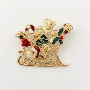 May include: A gold-tone metal brooch shaped like a Christmas sleigh. The design features a teddy bear, a Santa Claus figure, a candy cane, and holly leaves with red and green enamel accents. The sleigh has an openwork frame and a polished finish.