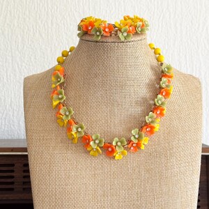 May include: A necklace and matching bracelet set featuring small, five-petaled flowers in shades of orange, yellow, and sage green. Each flower is accented with a tiny, clear, central rhinestone. The floral elements are linked together by a gold-tone metal chain.