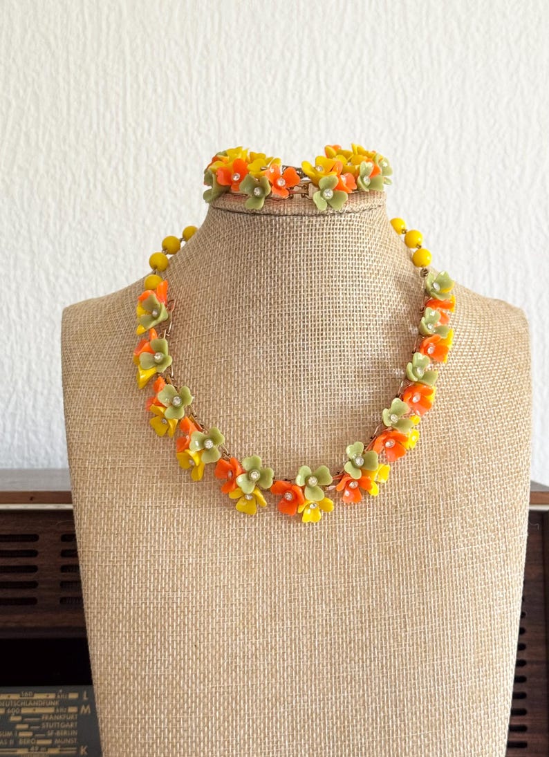 May include: A necklace and matching bracelet set featuring small, five-petaled flowers in shades of orange, yellow, and sage green. Each flower is accented with a tiny, clear, central rhinestone. The floral elements are linked together by a gold-tone metal chain.