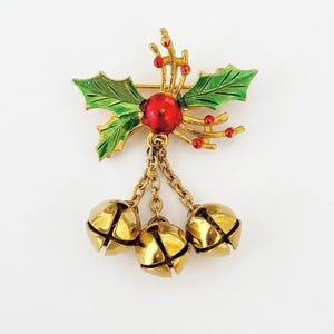 May include: A gold-tone brooch featuring green enamelled holly leaves and a central red berry. Three small gold-tone jingle bells hang from the holly base by short metal chains. The design is metallic with a polished finish and vibrant enamel accents.
