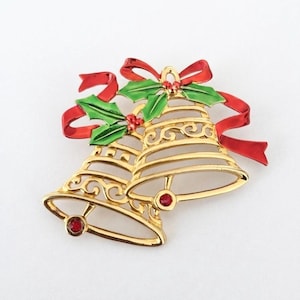 Pode incluir: Um broche de metal dourado em forma de dois sinos com detalhes recortados. Os sinos são decorados com folhas de azevinho em esmalte verde, bagas em esmalte vermelho e um laço de esmalte vermelho. Cada sino possui um pequeno strass vermelho redondo.
