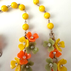 May include: A necklace featuring a strand of bright yellow beads connected to a cluster of orange, yellow, and sage green floral charms. Each flower is accented with a small, clear, round rhinestone at its center, linked by thin metal chains.