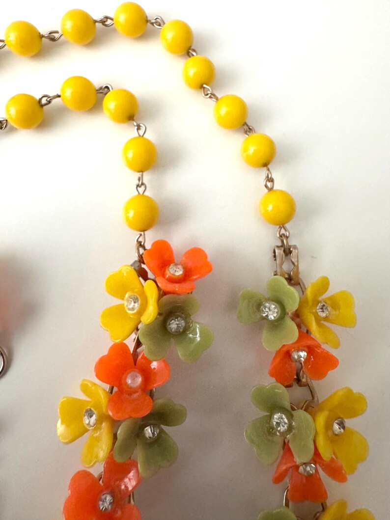 May include: A necklace featuring a strand of bright yellow beads connected to a cluster of orange, yellow, and sage green floral charms. Each flower is accented with a small, clear, round rhinestone at its center, linked by thin metal chains.