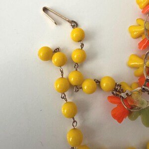 May include: A necklace featuring a series of round, glossy yellow beads linked by thin metal wire. The necklace includes a gold-toned metal hook clasp and decorative floral-shaped charms in vibrant shades of orange, yellow, and light green.