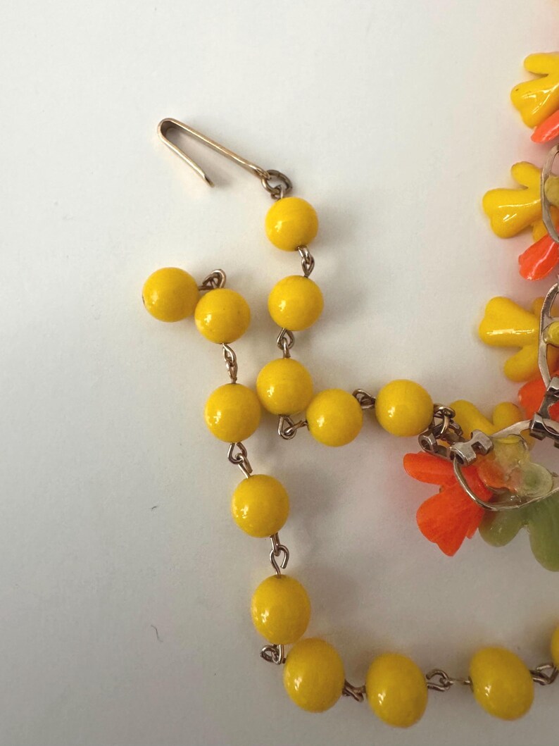 May include: A necklace featuring a series of round, glossy yellow beads linked by thin metal wire. The necklace includes a gold-toned metal hook clasp and decorative floral-shaped charms in vibrant shades of orange, yellow, and light green.