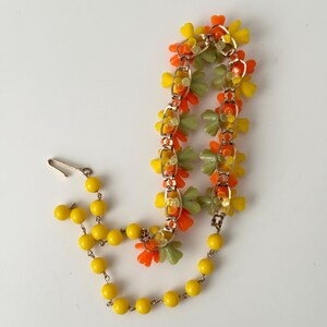 May include: A necklace featuring a gold-tone metal chain adorned with clusters of orange, yellow, and light green plastic flower-shaped beads, connected to a strand of round yellow beads with a metal hook clasp closure at the end.