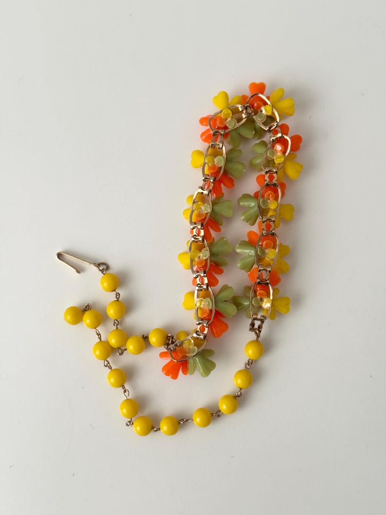 May include: A necklace featuring a gold-tone metal chain adorned with clusters of orange, yellow, and light green plastic flower-shaped beads, connected to a strand of round yellow beads with a metal hook clasp closure at the end.