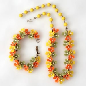 May include: A jewelry set consisting of a necklace and a bracelet featuring small orange, yellow, and sage green plastic flowers. Each flower has a clear rhinestone center. The necklace includes a strand of yellow beads and a metal hook clasp.