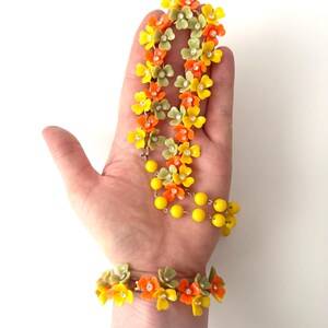 May include: A jewelry set consisting of a necklace and a bracelet featuring small orange, yellow, and sage green flower-shaped beads. Each flower has a tiny clear rhinestone at its center. The necklace includes a strand of bright yellow round beads.