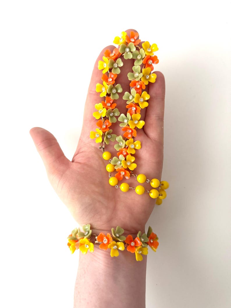 May include: A jewelry set consisting of a necklace and a bracelet featuring small orange, yellow, and sage green flower-shaped beads. Each flower has a tiny clear rhinestone at its center. The necklace includes a strand of bright yellow round beads.