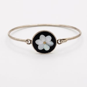 Peut inclure: Un bracelet jonc argenté doté d'une pièce centrale circulaire sur fond noir. Le centre présente une incrustation de fleur à six pétales en nacre blanche irisée avec un petit accent circulaire de couleur dorée au centre même.