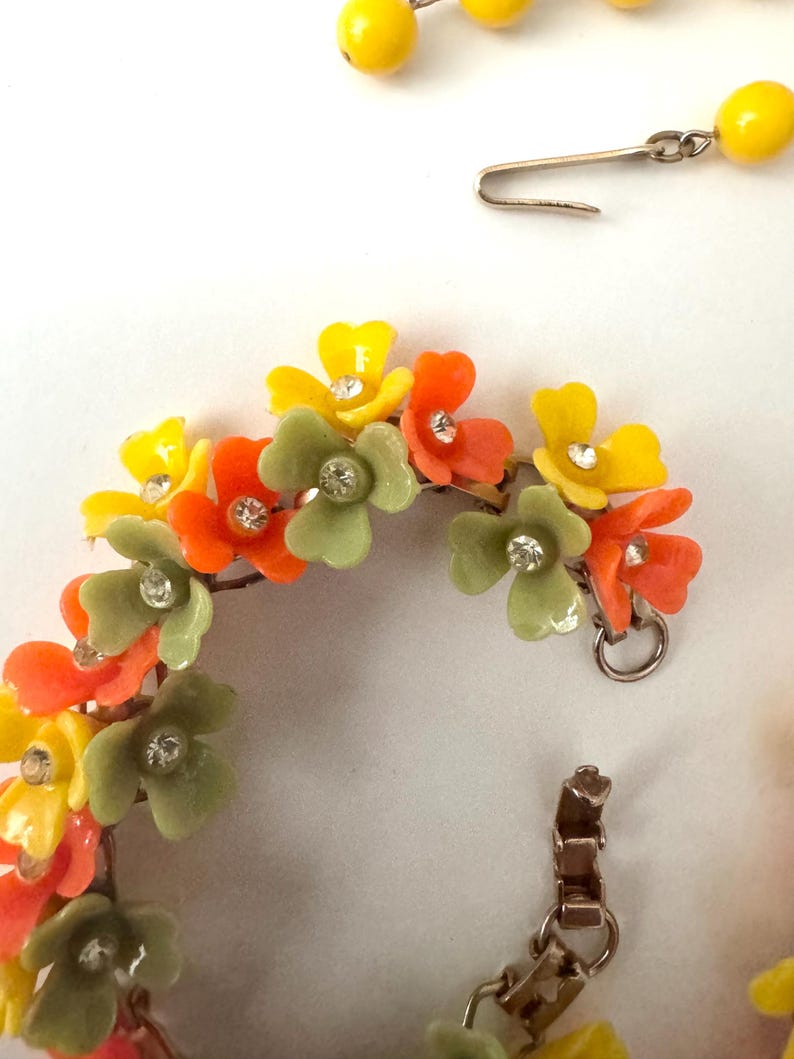 May include: A floral bracelet and matching earring set featuring yellow, orange, and sage green plastic flower charms. Each flower is accented with a small, clear, round rhinestone at the center. The bracelet has a metal clasp and link chain.