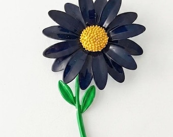 May include: A flower-shaped brooch featuring dark navy blue petals arranged in a circular pattern around a textured, bright yellow center. The brooch includes a slender, bright green stem with two small, pointed green leaves attached to the side.