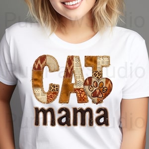 May include: White t-shirt with the words "CAT mama" in a patchwork design. The letters are in shades of brown and tan, with a heart-shaped paw print detail. The shirt is a casual style.