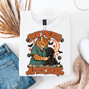Può includere: Una t-shirt bianca con la grafica di un gatto tigrato arancione che indossa una casacca verde acqua e usa uno stetoscopio su un cucciolo nero. Il testo "PAW PRINTS AFTER DARK" è stampato in arancione con pipistrelli e una luna.